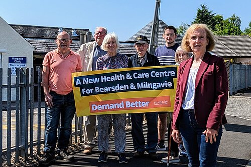 Susan Murray in front of a group of campaigners who are holding up a banner that reads "A new health centre for Bearsden and Milngavie. Demand Better. Scottish Liberal Democrats".