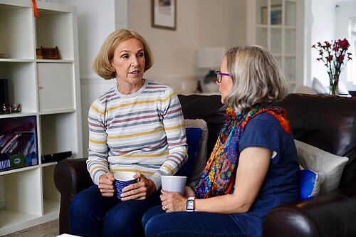 Susan Murray talking with resident on a couch, both holding coffee cups.