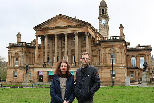 Anne Hannigan and Jack Clark standing next to each other, looking unhappy with Paisley Town Hall in the background.